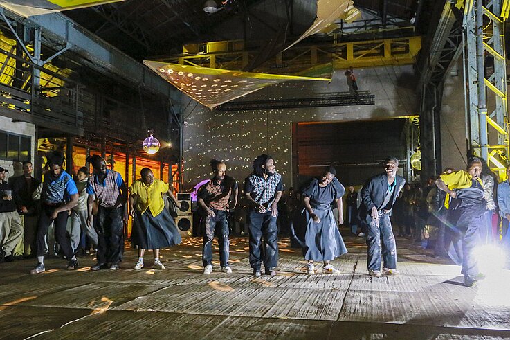 Eine Gruppe von Menschen tanzt energisch in einem großen Veranstaltungsort im Industriestil, der von bunten Lichtern und Discokugeln beleuchtet wird, mit Zuschauern im Hintergrund.