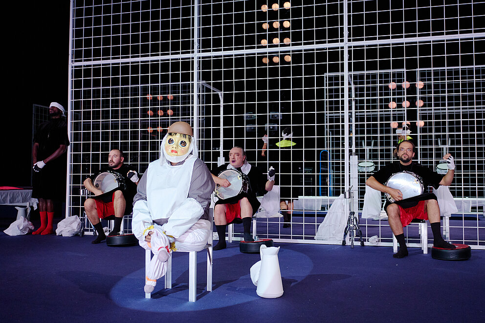 Auf dem Bild sieht man im Hintergrund eine Struktur aus weißen Eisengittern, im Vordergrund eine*n Schauspieler*in, der/die eine Puppenmaske trägt. Dahinter sind auch andere sitzende Schauspieler*innen zu erkennen.