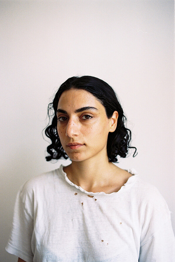 Portrait von Elvan Tekin in einem weißen T-Shirt und dunklen schulterlangen Locken