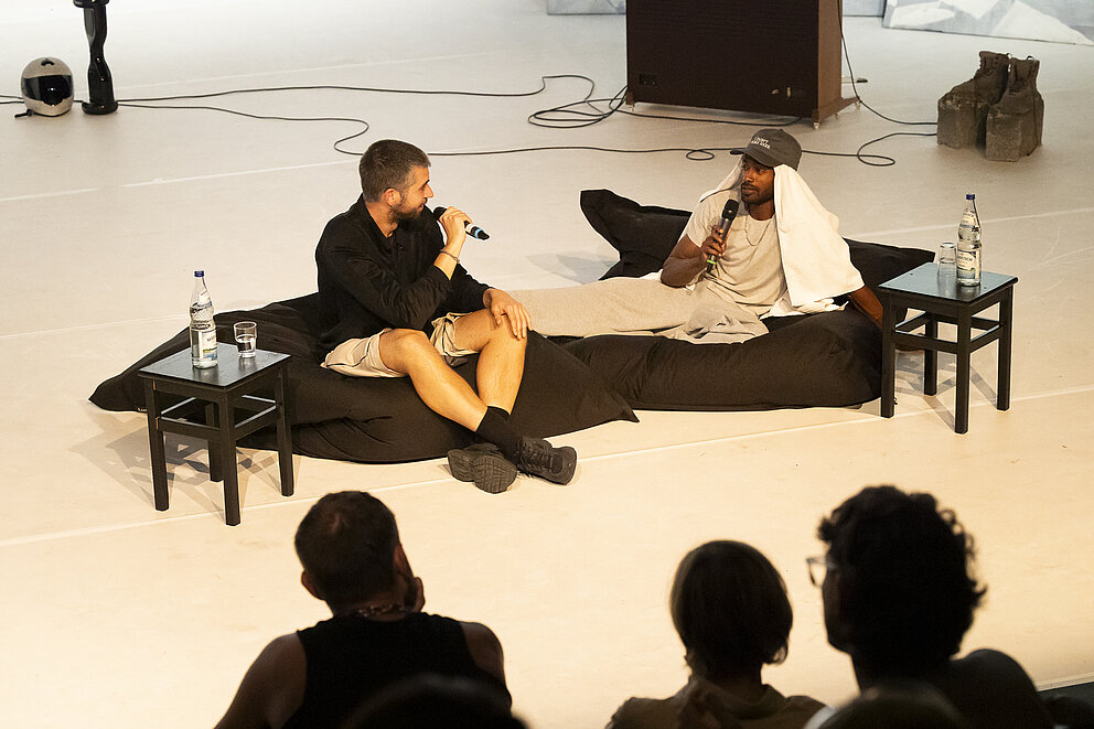 Auf dem Bild ist Beyond the Stage mit Jeremy Nedd und Mateusz Szymanówka zu sehen. Die beiden sitzen auf zwei Poufs und schauen sich an.