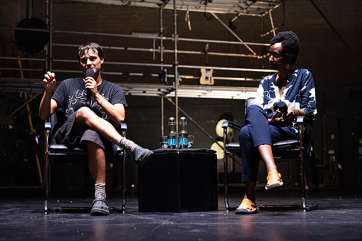 Nemó Flouret und Arlette-Louise Ndakoze stehen während Beyond the Stage auf der Bühne. Im Hintergrund des Bildes ist die Metallkonstruktion der Produktion zu sehen.