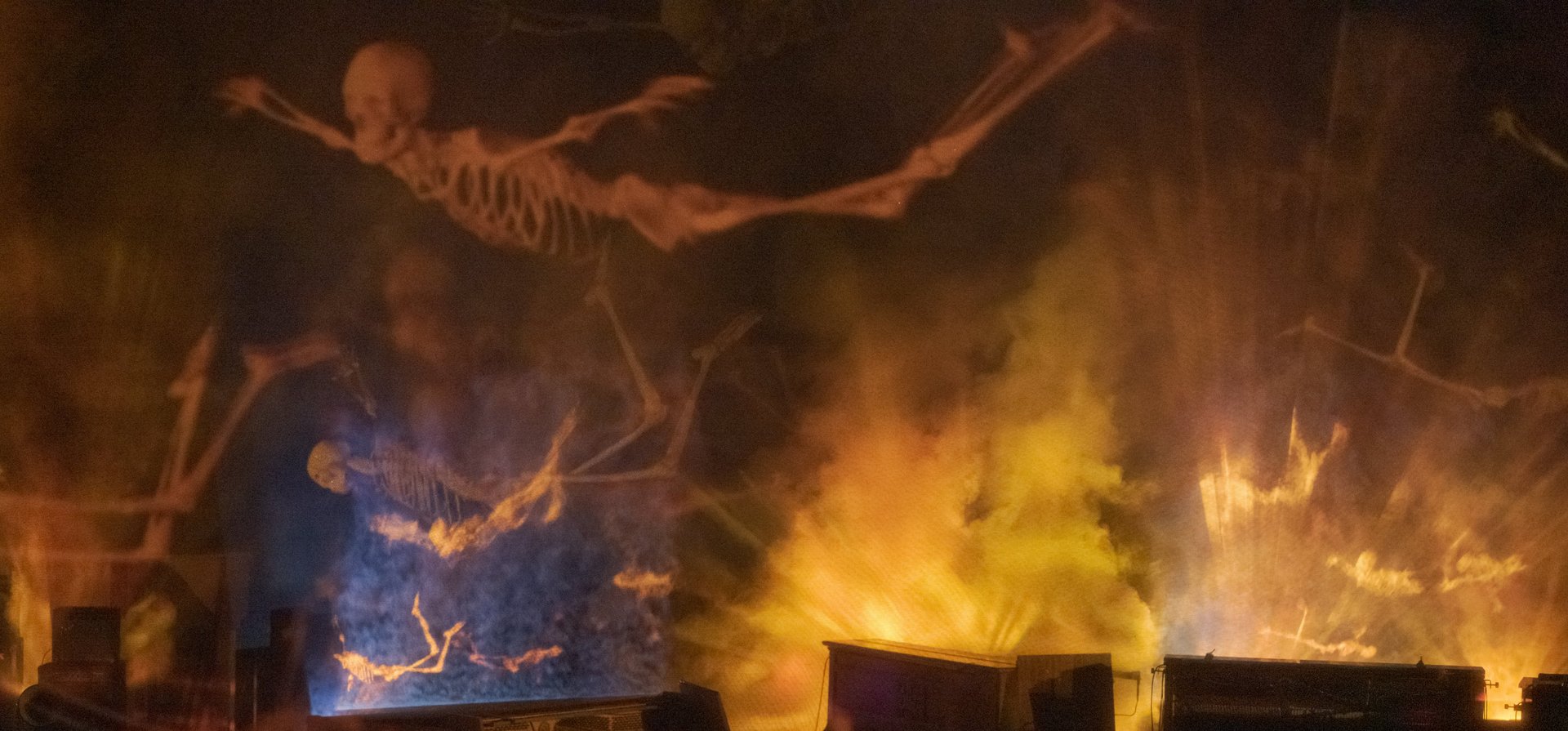 Photograph of a stage on which numerous upright pianos have an open case so that the hammer mechanism is free. Human skeletons hover above them. The foggy room makes the lighting appear dramatic.