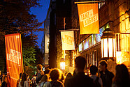 Auf dem Bild sind Menschen vor dem HAU1 nach der letzten Aufführung des Festivals zu sehen. Die Abendbeleuchtung ist eingeschaltet.