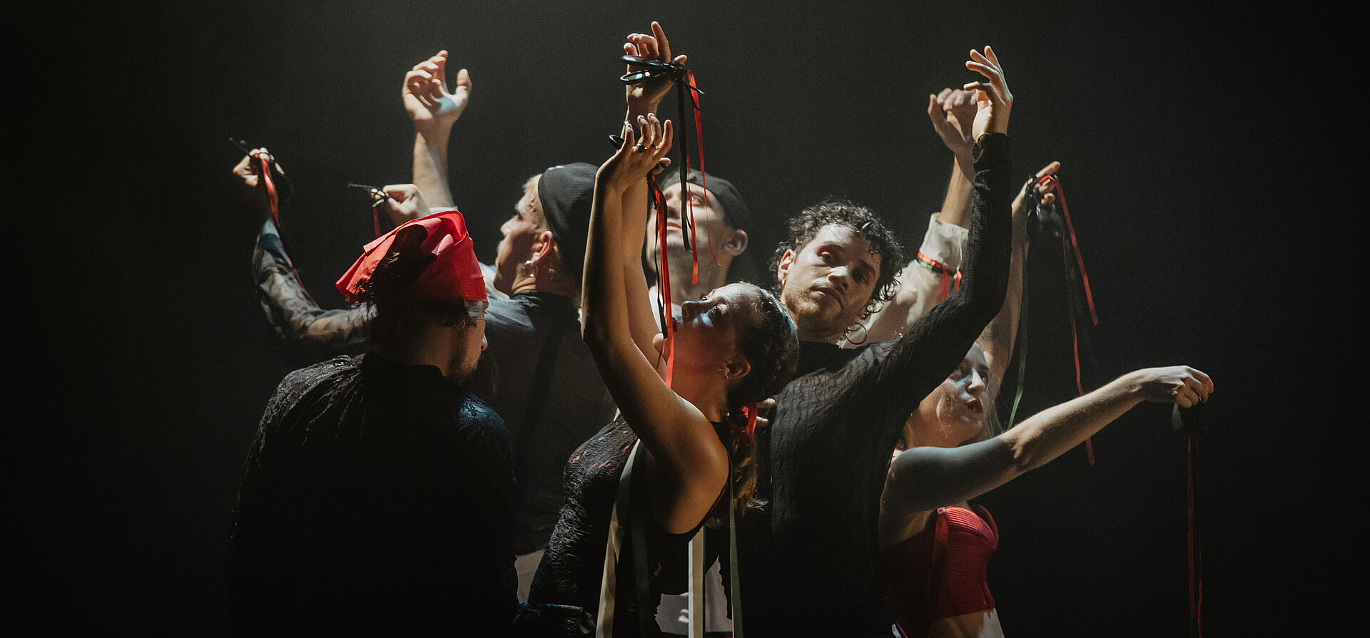 Acht Performer:innen stehen eng beisammen, die Arme anmutig erhoben, rote und grüne Bänder in den Händen – ein kraftvoller, ritueller Moment in dramatischem Bühnenlicht.
