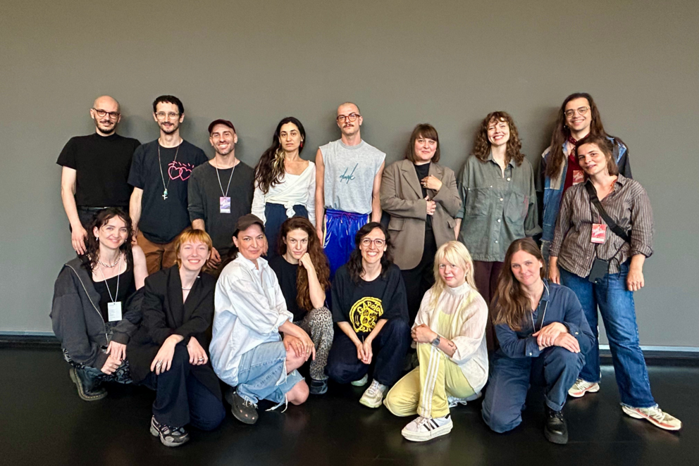 Auf dem Foto sind 16 Personen vor einer grauen Wand zu sehen, die sich für ein Gruppenfoto versammelt haben. Neben den 12 Künstler*innen sind auch 4 Personen aus dem Team von Tanz im August und HAU zu sehen.