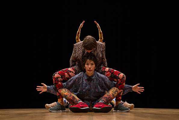 Drei Performer:innen in farbenfrohen, floralen und ornamentalen Outfits verschmelzen zu einer symmetrischen Figur auf der Bühne, vor schwarzem Hintergrund.