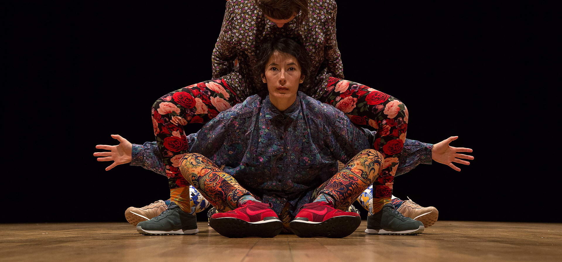 Drei Performer:innen in farbenfrohen, floralen und ornamentalen Outfits verschmelzen zu einer symmetrischen Figur auf der Bühne, vor schwarzem Hintergrund.