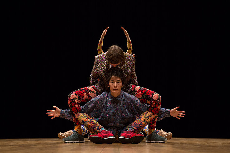 Drei Performer:innen in farbenfrohen, floralen und ornamentalen Outfits verschmelzen zu einer symmetrischen Figur auf der Bühne, vor schwarzem Hintergrund.