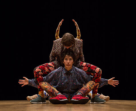Drei Performer:innen in farbenfrohen, floralen und ornamentalen Outfits verschmelzen zu einer symmetrischen Figur auf der Bühne, vor schwarzem Hintergrund.