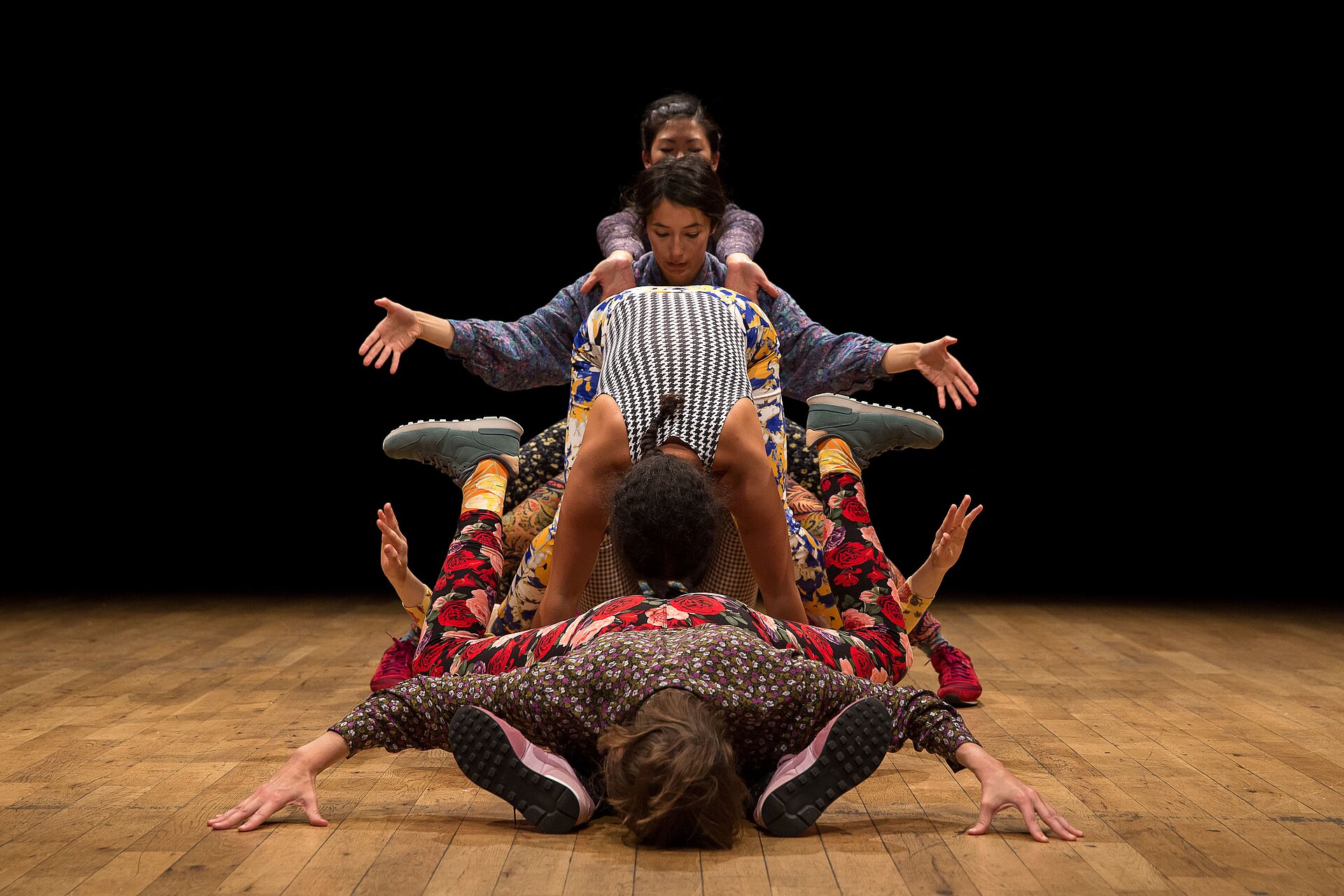 Sechs Performer:innen in farbenfroher Kleidung bilden eine übereinandergestapelte Formation auf der Bühne – eine dynamische, skulpturale Choreografie.