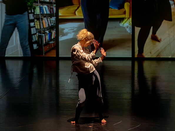 Eine barfüßige Performer:in mit blondem Haar tanzt allein auf dunkler Bühne, im Hintergrund sind großformatige Videoausschnitte mit Innenraumszenen zu sehen.