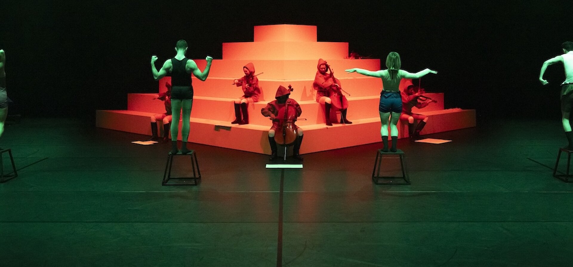 Performer:innen auf Hockern und Musiker:innen in roten Kapuzen posieren vor einer stufenförmigen weißen Plattform im grünen und roten Licht.