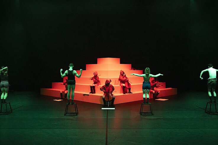 Performer:innen auf Hockern und Musiker:innen in roten Kapuzen posieren vor einer stufenförmigen weißen Plattform im grünen und roten Licht.