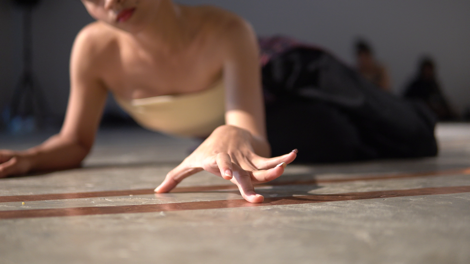 Performer:in liegt am Boden, Arm ausgestreckt, zwei Finger berühren beinahe den Boden – Moment körperlicher Spannung und Ruhe.